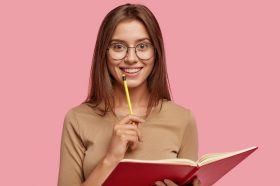Photo of happy brunette woman with positive smile, carries textbook, holds pencil for writing, makes notes while listens some information, poses over pink background. Its time for creating new poem!