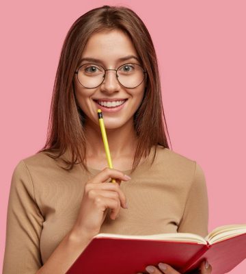 Photo of happy brunette woman with positive smile, carries textbook, holds pencil for writing, makes notes while listens some information, poses over pink background. Its time for creating new poem!