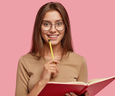 Photo of happy brunette woman with positive smile, carries textbook, holds pencil for writing, makes notes while listens some information, poses over pink background. Its time for creating new poem!